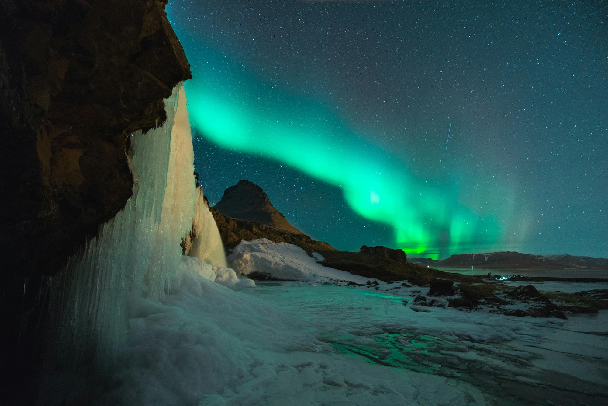 Mesmerizing display of the Northern Lights dancing across the night sky above a frozen waterfall and snow-covered landscape in Iceland, with Kirkjufell mountain in the background.